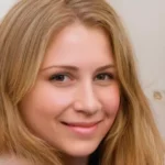 A photograph of a person's face. The person is looking directly at the camera and has a friendly smile. They have short dark hair and are wearing a white shirt or blouse. The background is plain and neutral, with no distinguishing features